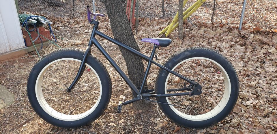 Black fat tire bicycle with purple handlebars and seat leaning against a tree.
