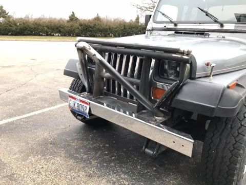 1992 Jeep Wrangler - A close-up, low-angle shot of the front bumper and grille of a silver Jeep Wrangler. The vehicle features a custom chrome bumper with a bru…
