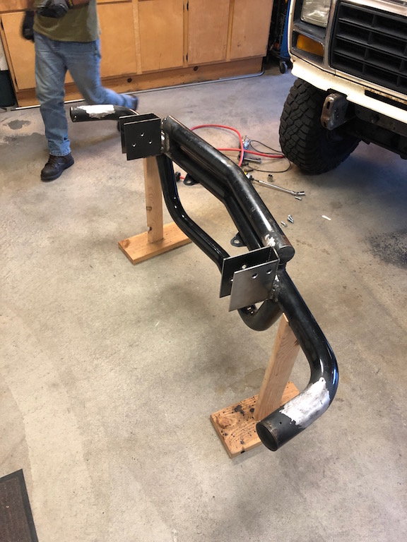 Custom tubular front bumper assembly on wooden stands in a garage. Part of a white vehicle is visible in the background