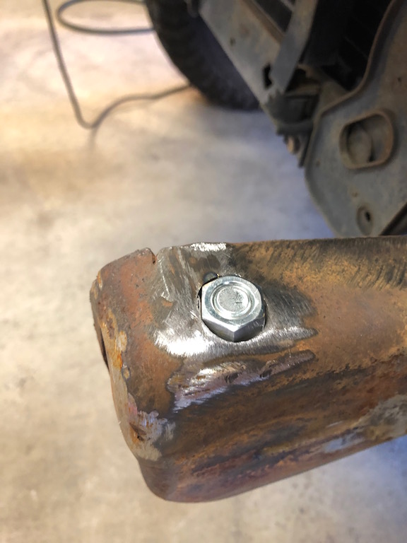 Rusty metal bracket with a new bolt and nut, showing weld marks and a tire in the background