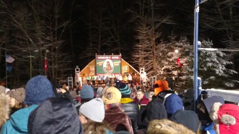 2004 Aston Martin Virage - An audience gathers at night to watch a performance on a stage, with snow-dusted trees in the background.