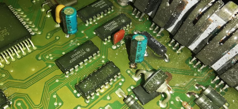 Close-up of a green circuit board with various electronic components like chips, capacitors, and transistors. Some components have visible part numbers