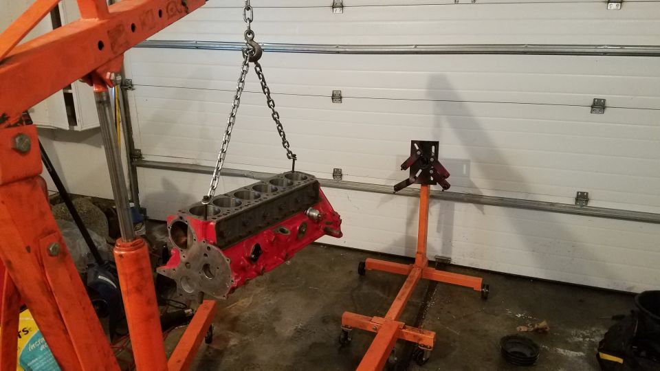 Red engine block suspended by chains from an orange engine hoist, with an engine stand nearby