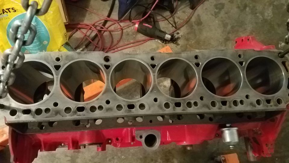 Engine block with six cylinders, suspended by a chain. Tools and a bag are in the background