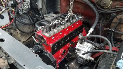 1985 Ford Bronco - A red V8 engine is being lowered into the engine bay of a vehicle with a gray fender and red firewall. The engine is partially assembled wi…