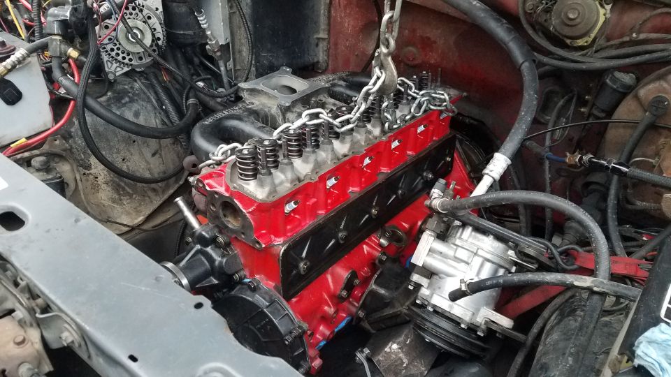 Red V8 engine being lowered into vehicle engine bay with chain hoist.