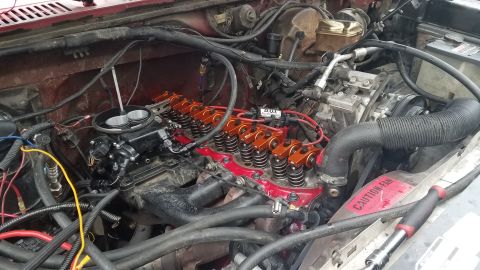 1985 Ford Bronco - Close-up view of the engine bay of a red Ford Bronco, focusing on the engine with orange roller rockers and a black carburetor.