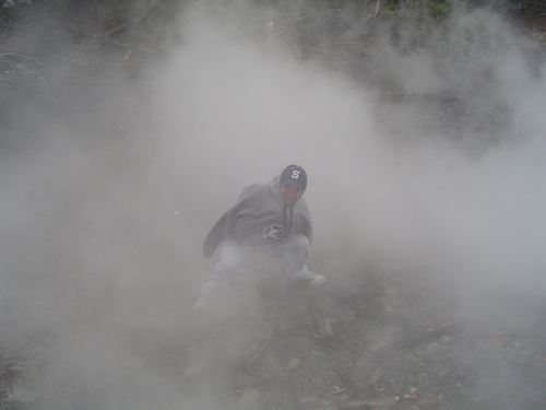 guy in a hoodie and baseball cap crouched down in thick smoke, with a Supermotors logo at the bottom right