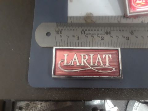 1994 Ford Bronco - A chrome and red emblem that reads "LARIAT" is shown next to a metal ruler. The emblem appears to be a vehicle part.