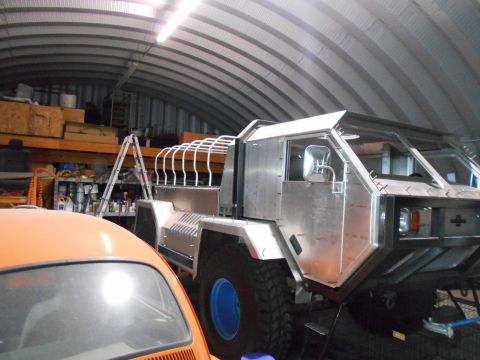 1992 Ford Bronco - A custom-built, unfinished vehicle with a silver metal body is parked inside a metal Quonset hut garage. An orange car is partially visible…