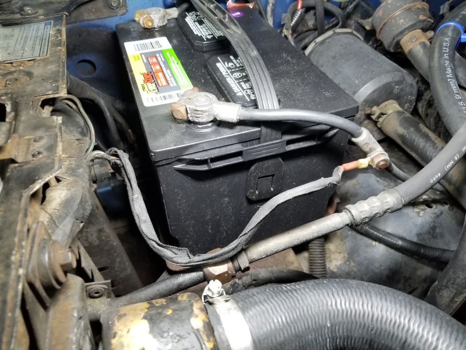 car battery with terminals and cables, surrounded by engine hoses and metal parts