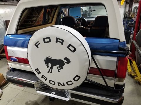 1989 Ford Bronco - The rear of a blue and white Ford Bronco is shown, featuring a white spare tire cover with the Ford Bronco logo and a horse emblem. The veh…