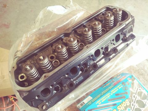 1996 Ford Bronco - A black engine cylinder head with multiple valve springs and retainers is shown partially covered by plastic packaging. A blue box of "Bolt…
