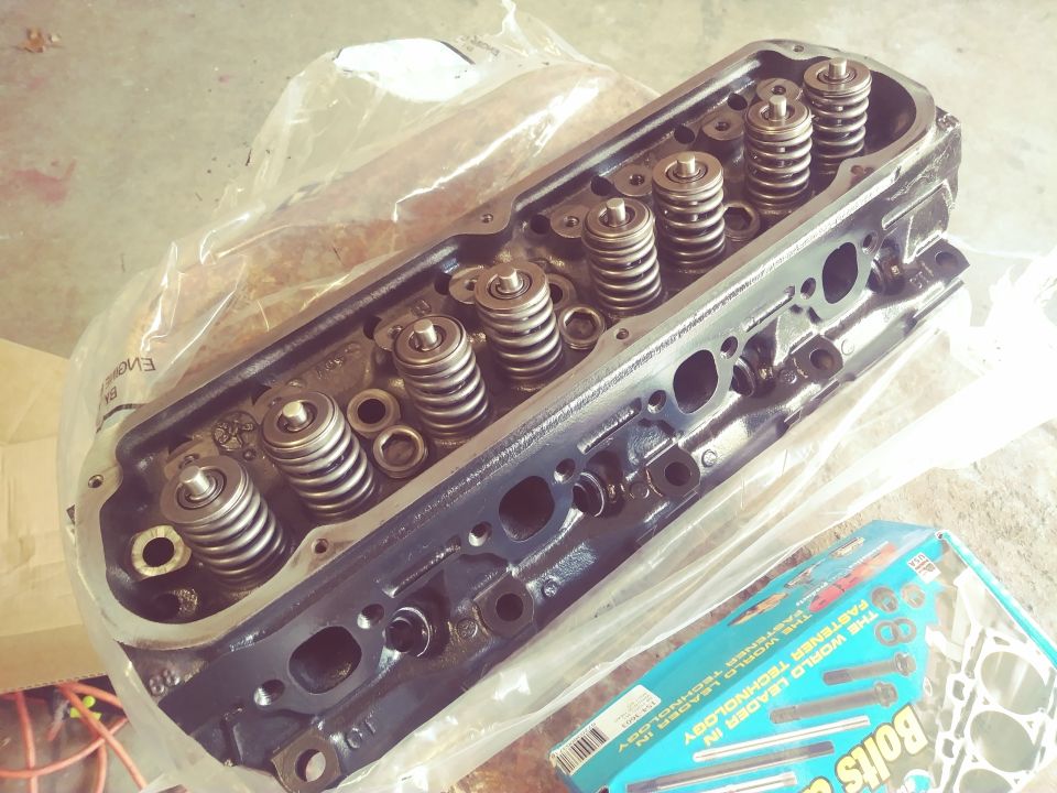 Black engine cylinder head with valve springs, partially covered in plastic, next to a box of bolts.
