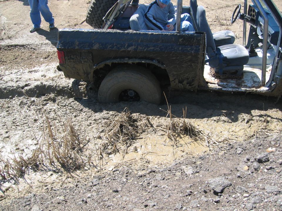Mud-covered off-road vehicle with one wheel deep in mud, two people in the cab