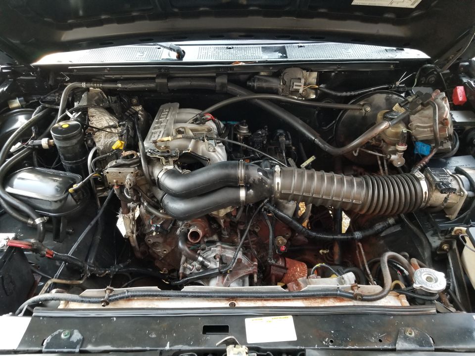 Engine bay of a black Ford Bronco showing inline-six EFI engine, intake hoses, and brake booster