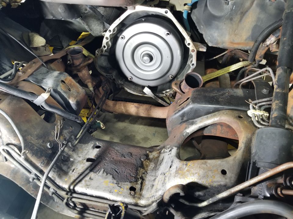 Transmission torque converter housing bolted to engine block, with rusty frame rails and exhaust pipes visible