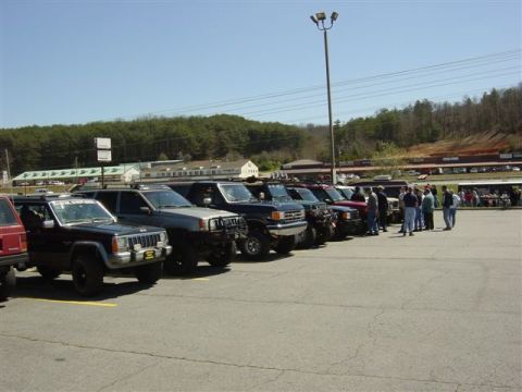 1995 Ford Bronco - Row of lifted 4x4s lined up in a parking lot at a trail-ride staging area, led by a dark blue Jeep Cherokee XJ with a tubular front bumper,…