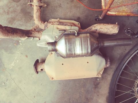 1996 Ford Bronco - A catalytic converter and exhaust pipe assembly is laid out on a concrete floor. The parts appear to be heavily rusted and covered in dirt.