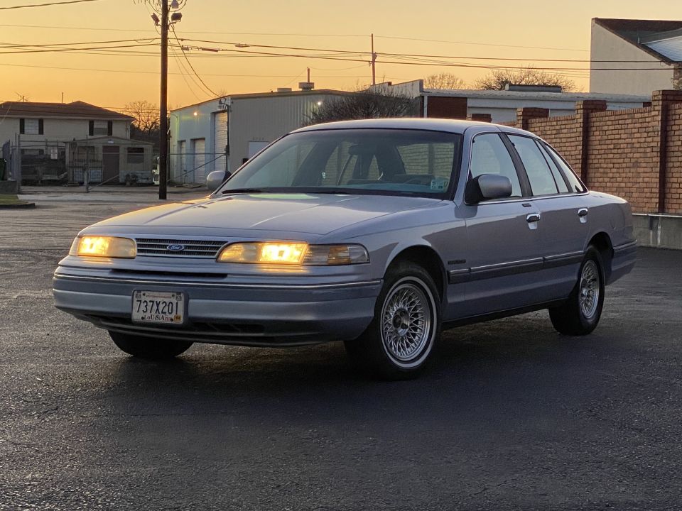 1994 Ford Crown Victoria sedan parked on asphalt lot at sunset, headlights on, Louisiana license plate visible.