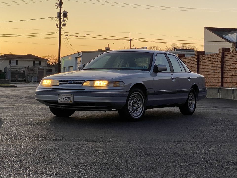 1994 Ford Crown Victoria sedan, silver, parked with headlights on at sunset.