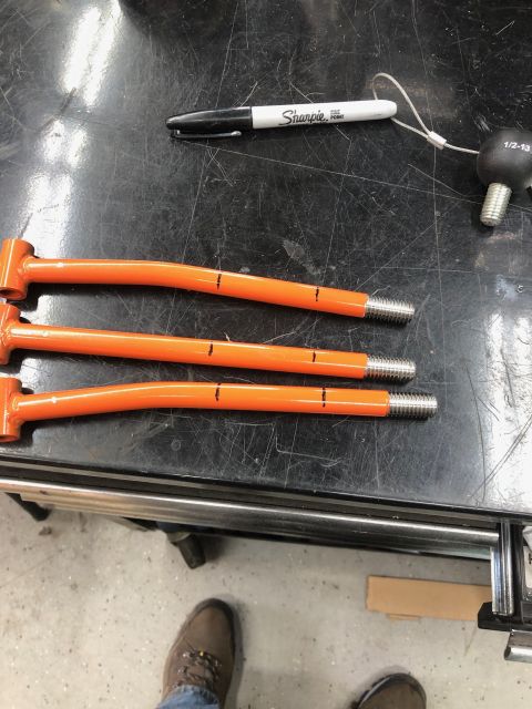 1992 Jeep Wrangler - Three orange, curved metal rods with threaded ends are laid out on a black workbench. Each rod has three black marks indicating measurement…