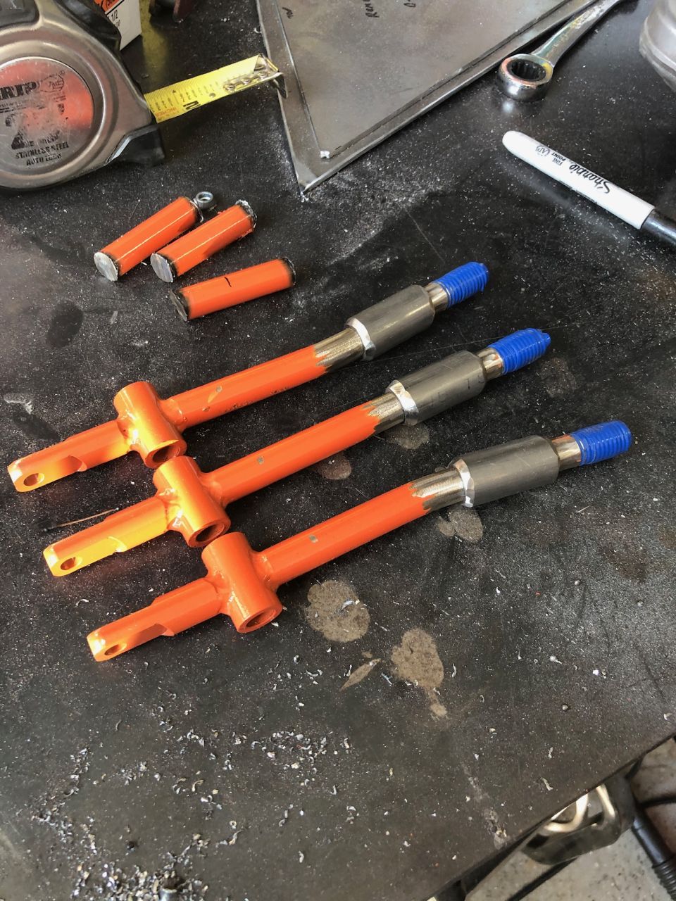 Three orange metal parts with silver and blue ends on a workbench.
