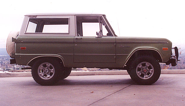 Green vintage Ford Bronco with white hardtop, side view, parked on concrete overlook