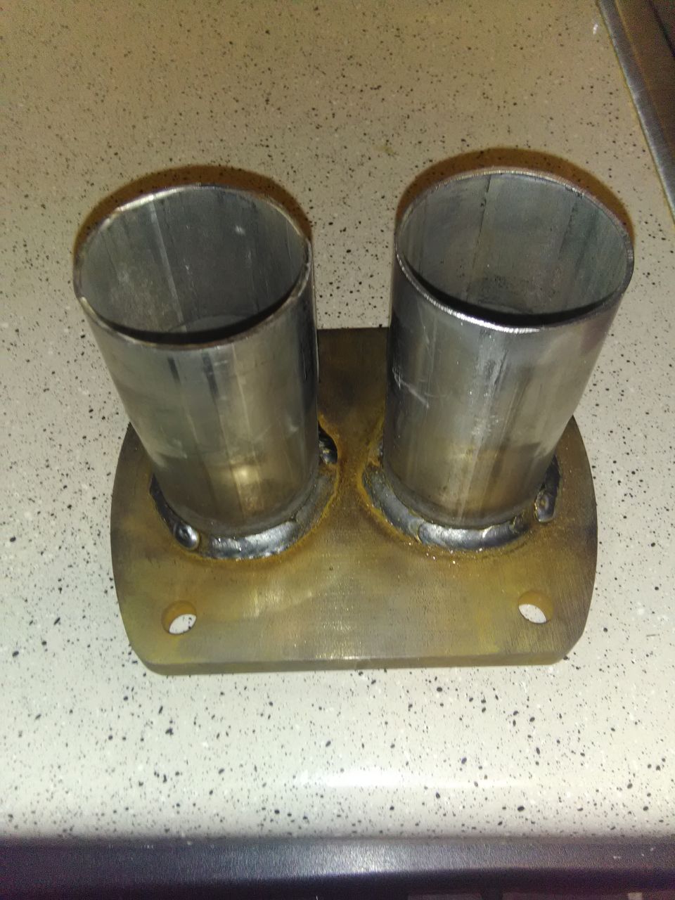 Two steel pipes welded to a flat flange plate with bolt holes, showing weld beads and heat discoloration