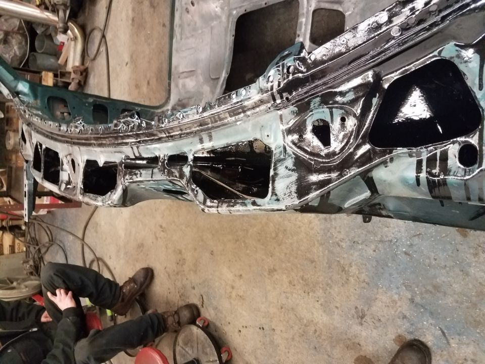 Vehicle frame with black coating, two people working nearby.