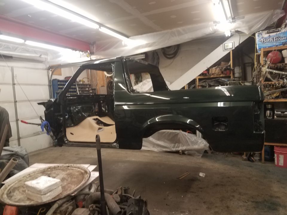 Dark green Ford Bronco body suspended in a garage, doors open, undergoing restoration.