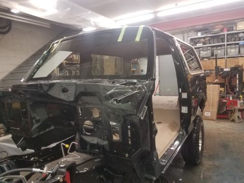 1994 Ford Bronco - The black body of a Ford Bronco is shown in a workshop setting, with the windshield taped in place and the interior partially visible. The…