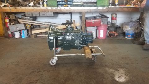 1994 Ford Bronco - A dark green, heavy-duty transmission is mounted on a rolling cart in a workshop. The transmission has a manual shifter and appears to be a…