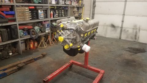 1994 Ford Bronco - A V8 engine block, covered in yellow protective caps, is mounted on a red engine stand in a garage. Shelving units filled with tools and su…