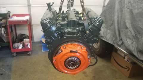 1994 Ford Bronco - A dark green V8 engine with an orange clutch assembly is suspended by chains in a garage setting. A red tool cart is visible to the left, a…