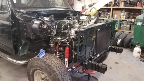 1994 Ford Bronco - The front end of a dark green 1994 Ford Bronco is shown disassembled in a garage setting. The engine, radiator, and suspension components a…