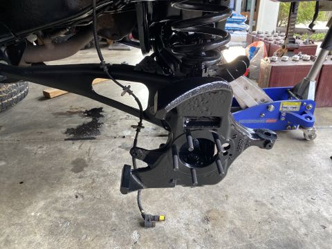 1994 Ford Bronco - A black painted suspension part, likely a steering knuckle, is shown attached to a control arm. A blue hydraulic jack is visible in the bac…