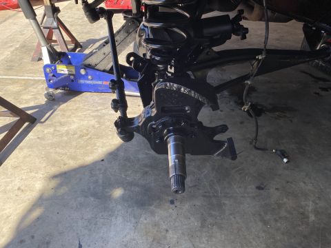 1994 Ford Bronco - A black painted suspension component, likely a steering knuckle and hub assembly, is shown with a blue hydraulic jack and jack stands in th…