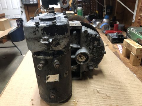 1980 Ford Bronco - A close-up, low-angle view of a vintage starter motor or generator with a transfer case attached, sitting on a workbench. The metallic gray…