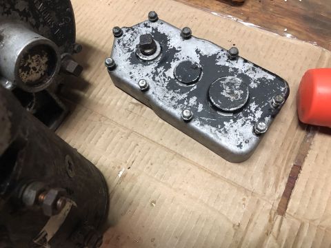 1980 Ford Bronco - A close-up view of a metal gearbox cover with a black painted surface that is chipped and worn, revealing silver metal underneath. Several…