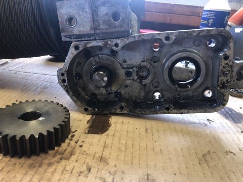 1980 Ford Bronco - A disassembled metal gearbox with a large metal gear in the foreground, resting on a cardboard surface with oil stains.