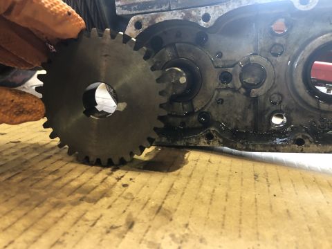 1980 Ford Bronco - A gloved hand holds a metal gear with visible teeth next to a greasy, disassembled engine block with circular openings and bolt holes.