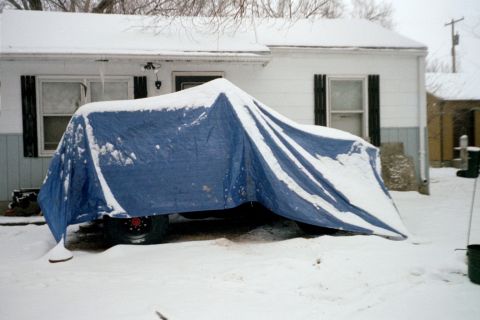 1979 Ford Bronco - A vehicle is covered by a blue tarp and snow in a residential driveway during winter.