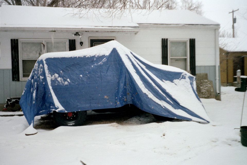 Vehicle covered by blue tarp in snow, parked in front of a house