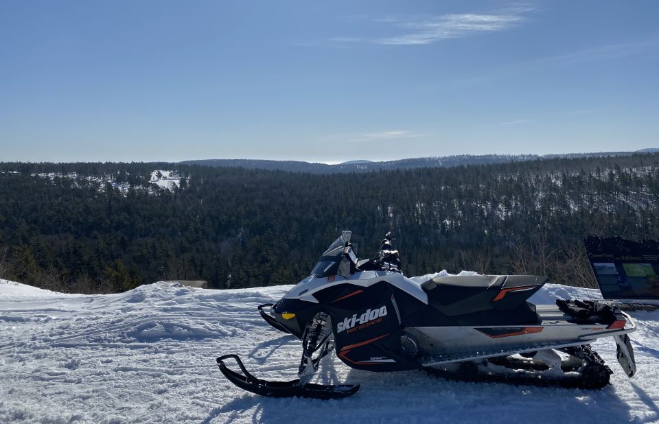 Ski-Doo snowmobile parked on a snowy overlook with forest and hills in the background.