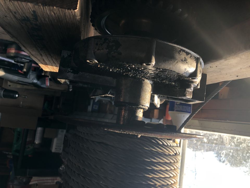 Winch drum with cable and metal housing mounted under a wooden beam.