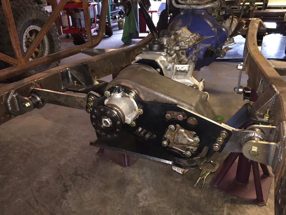 Vehicle chassis with transmission and transfer case, supported by jack stands. A large tire is visible on the left