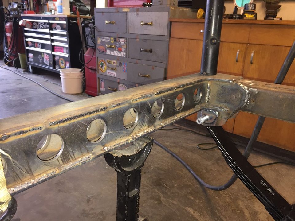 Custom steel frame with circular cutouts and welds, supported by jack stands, with a leaf spring visible.