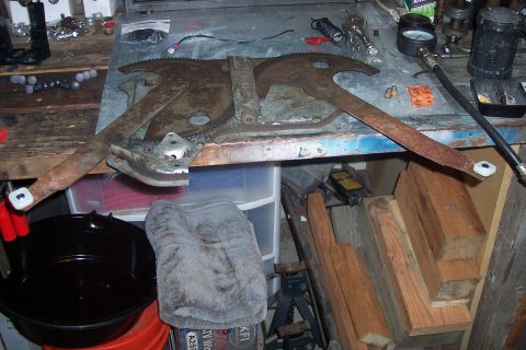 1995 Ford Bronco - A close-up of a rusty window regulator mechanism, likely for a vehicle door, resting on a cluttered workbench.