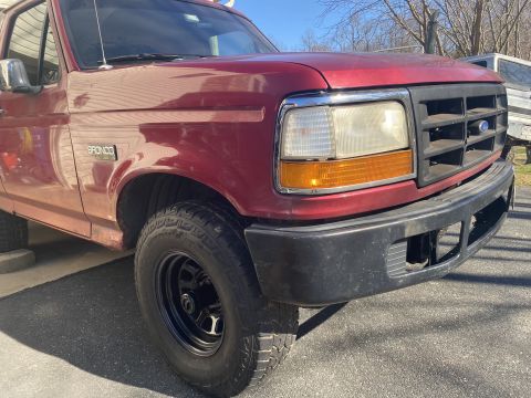 1994 Ford Bronco - A close-up, low-angle shot of the front passenger side of a red Ford Bronco. The vehicle is parked outdoors on an asphalt surface.